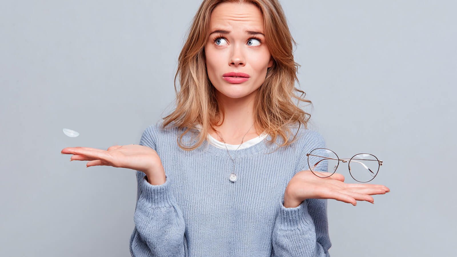 A young woman with a confused expression holds a single contact lens in one hand and a pair of glasses in the other, symbolizing the common question about the differences between eyeglass and contact lens prescriptions.