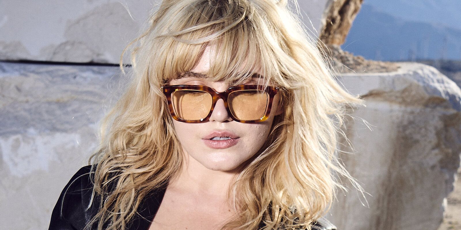 A close-up of a woman in a casual top styling retro-inspired sunglasses with faint yellow-tinted lenses.