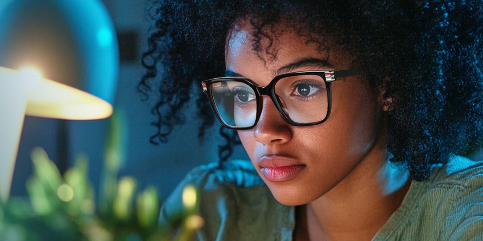 Focused young woman with curly hair and black-rimmed eyeglasses working at a desk under a desk lamp, with a plant in the foreground.