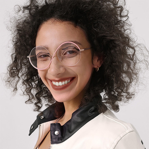 A smiling woman with voluminous curly dark hair wearing unique pink round asymmetrical glasses and small gold hoop earrings, demonstrating hairstyles that flatter frames.
