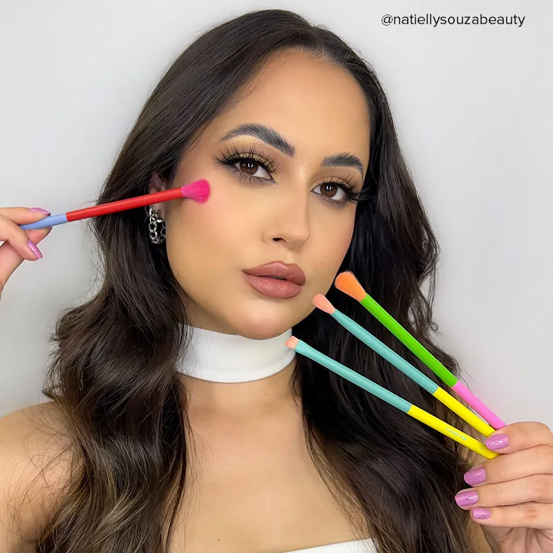 A close-up of a woman applying blush with a colorful brush, showcasing bold gold eyeshadow and winged eyeliner designed to pop behind glasses frames.