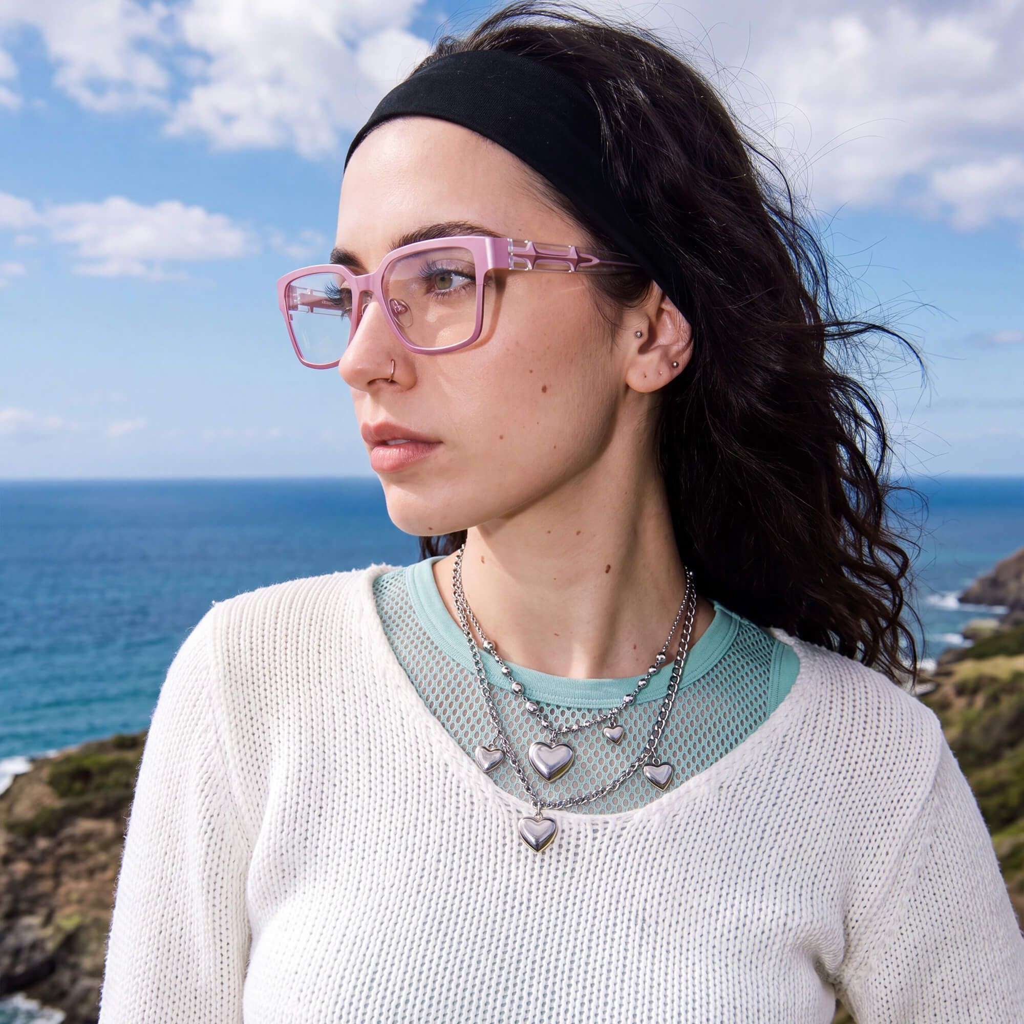 A young woman with long dark hair and a headband posing outdoors by the sea, wearing stylish pastel pink rectangular glasses with detailed temples.