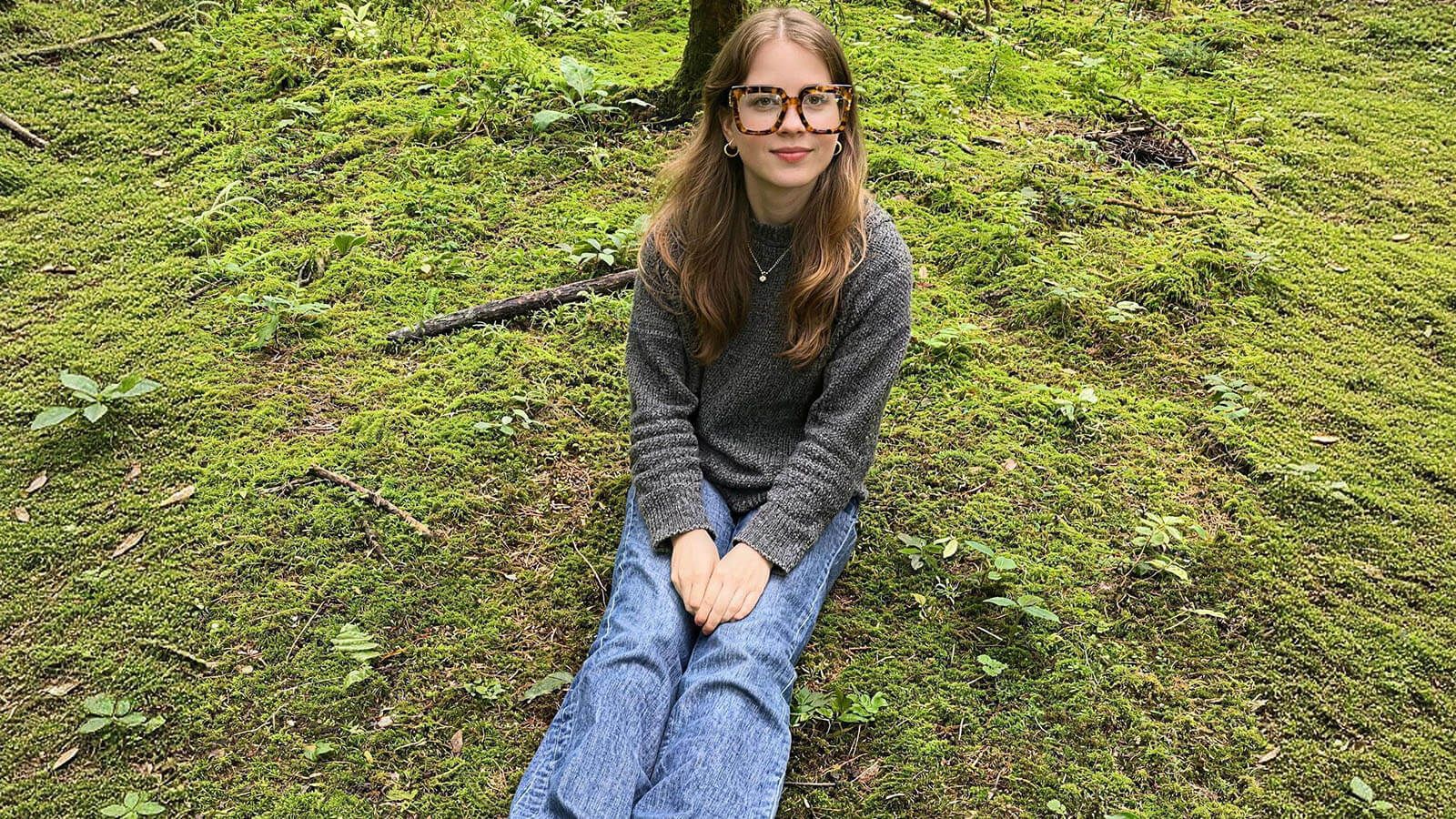 Young girl with long hair and tortoiseshell eco glasses sitting on a grassy forest floor, wearing a gray sweater and blue jeans